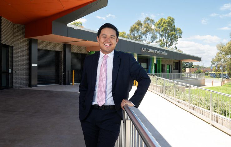 A smiling man leaning on a metal bannister in front of a large sports pavilion.