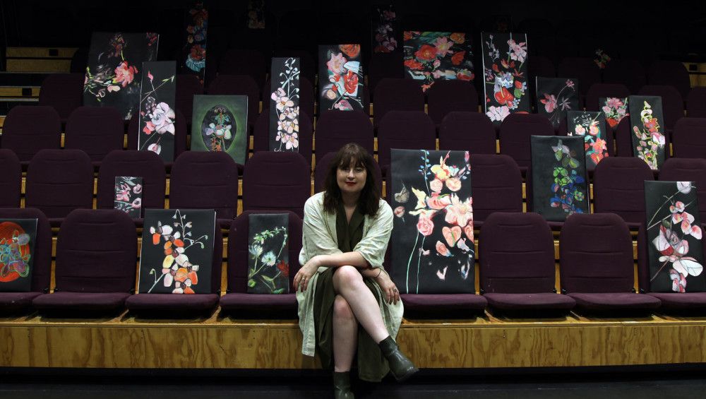 Artist Josephine Mead sitting in front of floral paintings positioned in theatre seats. 