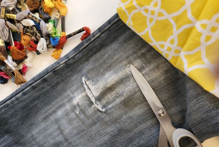 Ripped blue jeans with scissors. A piece of yellow and white patterned fabric is in the right top corner. A pile of embroidery threads are in the left top corner. 
