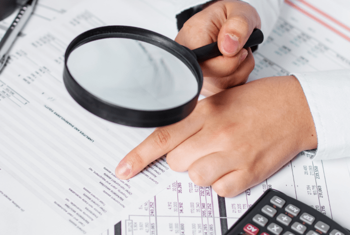 A business person using a magnifying glass and calculator to audit documents.