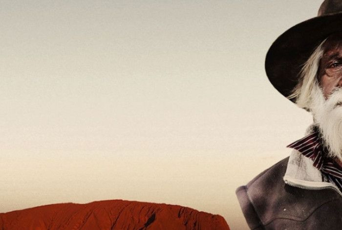 A senior first nation's man wearing a hat and sporting a long beard stands in front Uluru embodying a sense of history and character.