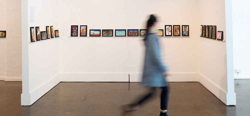 A blurred figure walks in front of a wall of 9 by 5 artworks.