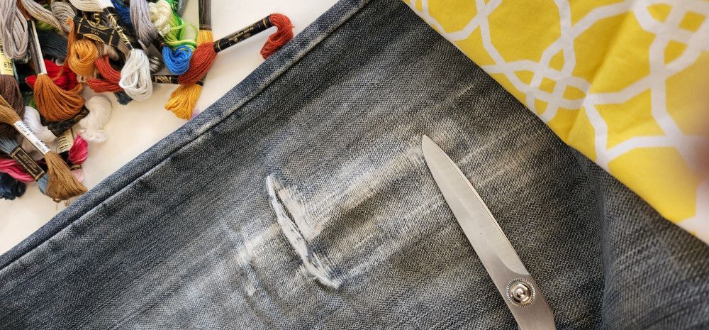 Ripped blue jeans with scissors. A piece of yellow and white patterned fabric is in the right top corner. A pile of embroidery threads are in the left top corner. 
