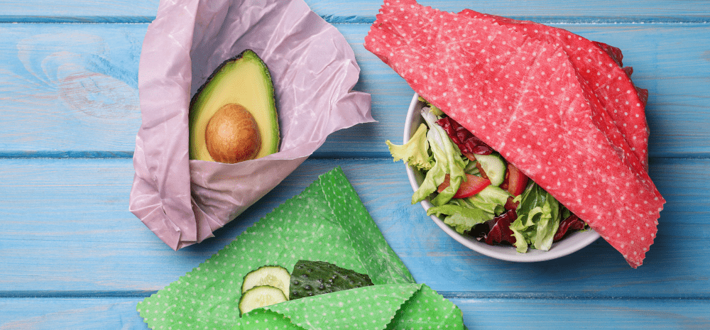 An avocado, a cucumber and a bowl of salad in colourful beeswax wraps on a blue background.