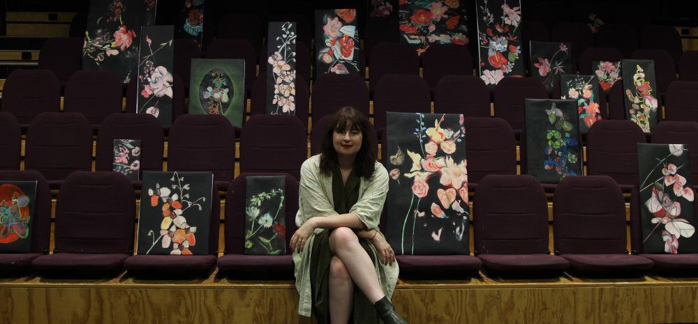 Artist Josephine Mead sitting on theatre seats with their paintings.