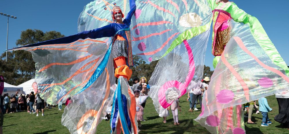 Two characters on stilts with flowing fairy wings walking through a park on a blue sky day.