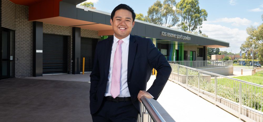 A smiling man leaning on a metal bannister in front of a large sports pavilion.