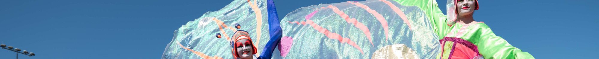 Two characters on stilts with flowing fairy wings walking through a park on a blue sky day.