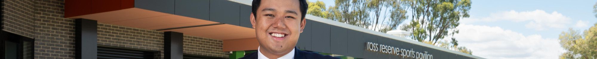 A smiling man leaning on a metal bannister in front of a large sports pavilion.