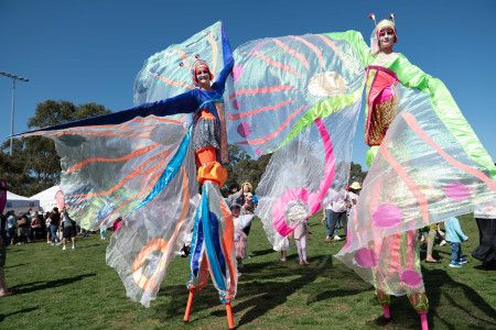 Two characters on stilts with flowing fairy wings walking through a park on a blue sky day.