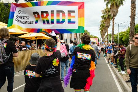 A group of people walking down a street lined by crowds under a banner that reads Greater Dandenong Pride